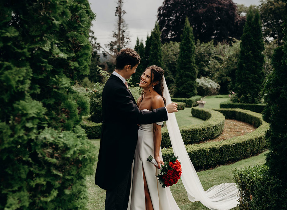 a smiling bride holding a bouquet of red roses with a groom in a lush green garden setting with manicured hedges at Achnagairn Castle