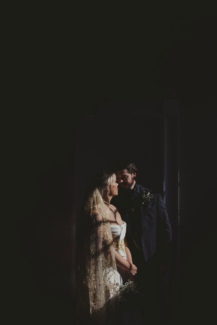 a bride and groom standing by a window with sunlight shining onto them.