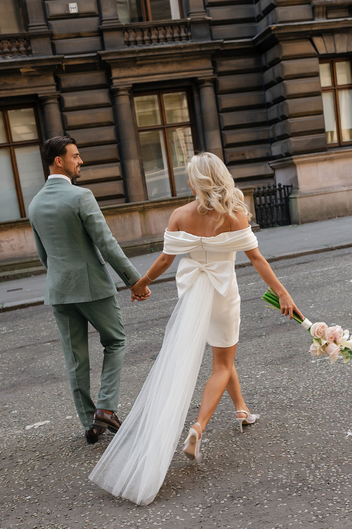 Bride and groom walk hand in hand through Glasgow streets, bride holding pale pink rose bouquet trailing a soft tulle train