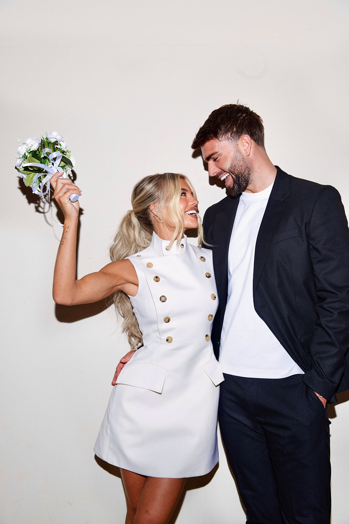 Bride in a modern high-neck mini wedding dress with gold button detailing holds a bouquet up playfully while smiling at her partner; he wears a relaxed black suit with white T-shirt against a plain background