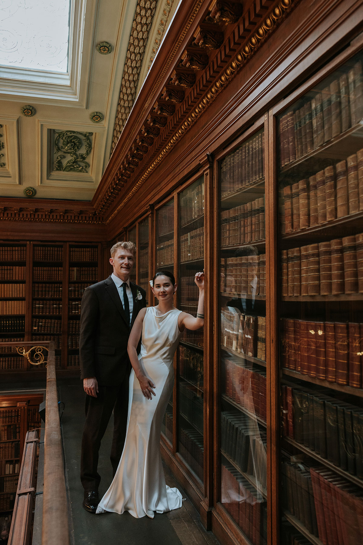 Bride and groom posing in front of wooden bookcases inside a grand library hall.