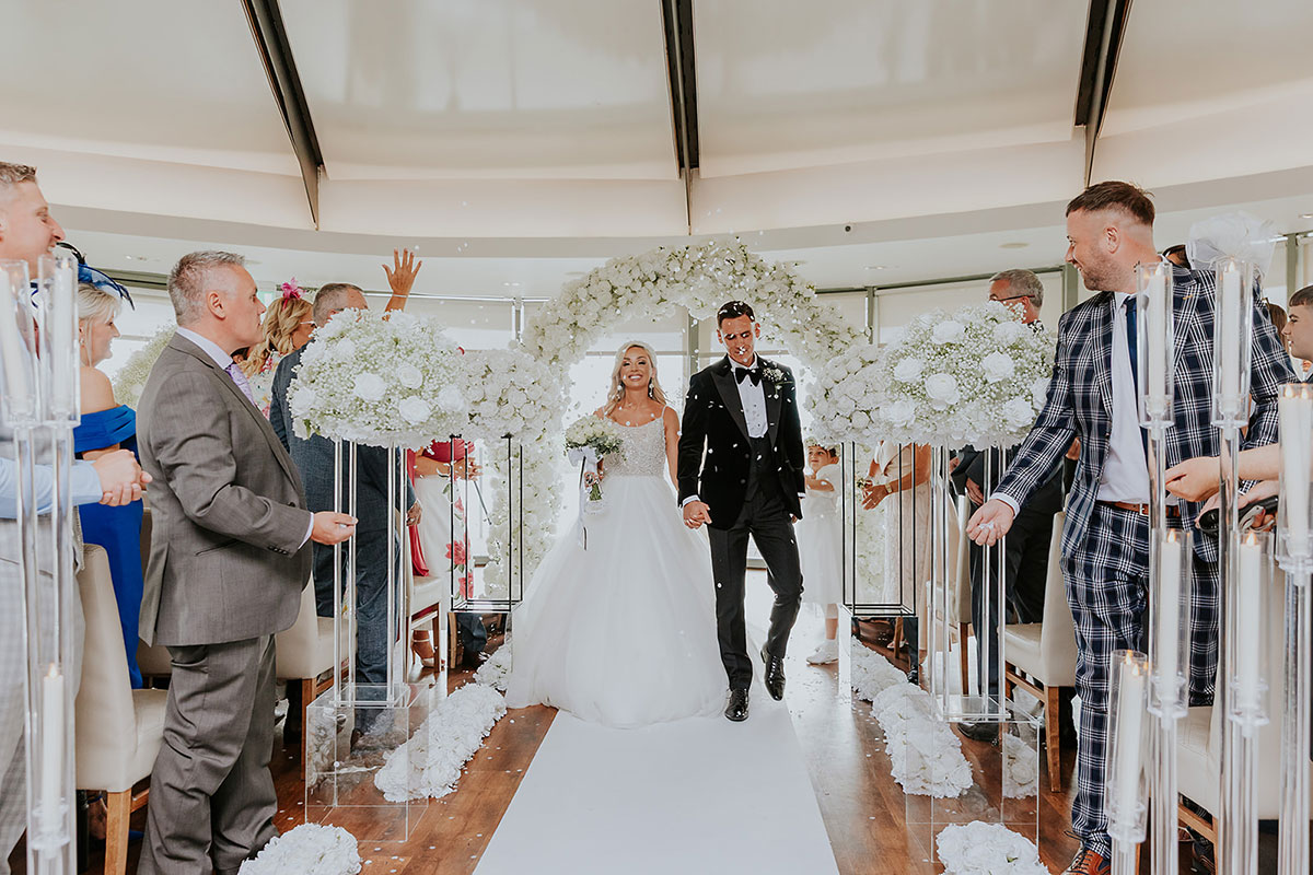 blonde bride in ballgown and groom in tux hold hands, walking up white aisle as friends and family throw white confetti at them