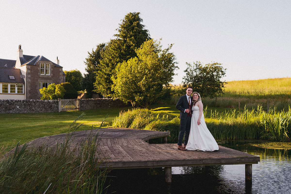 A bride and groom stand together on a wooden jetty overlooking a tranquil pond at sunset, with tall grasses, trees and a stone-built country house behind them. The warm golden light creates a peaceful rural setting