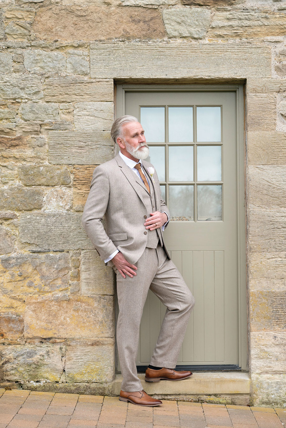 a man wearing a beige suit leaning against a pale green painted door