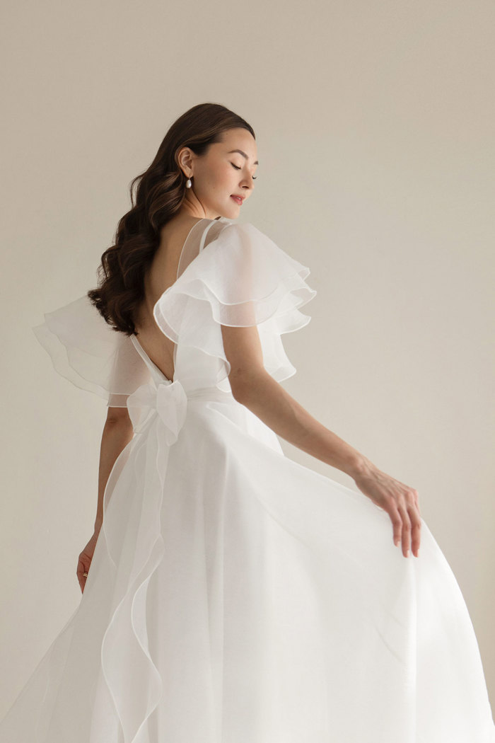 dark haired model wears floaty white wedding dress with sheer layer over bodice providing fluttery sleeves and a small bow at the back of waist