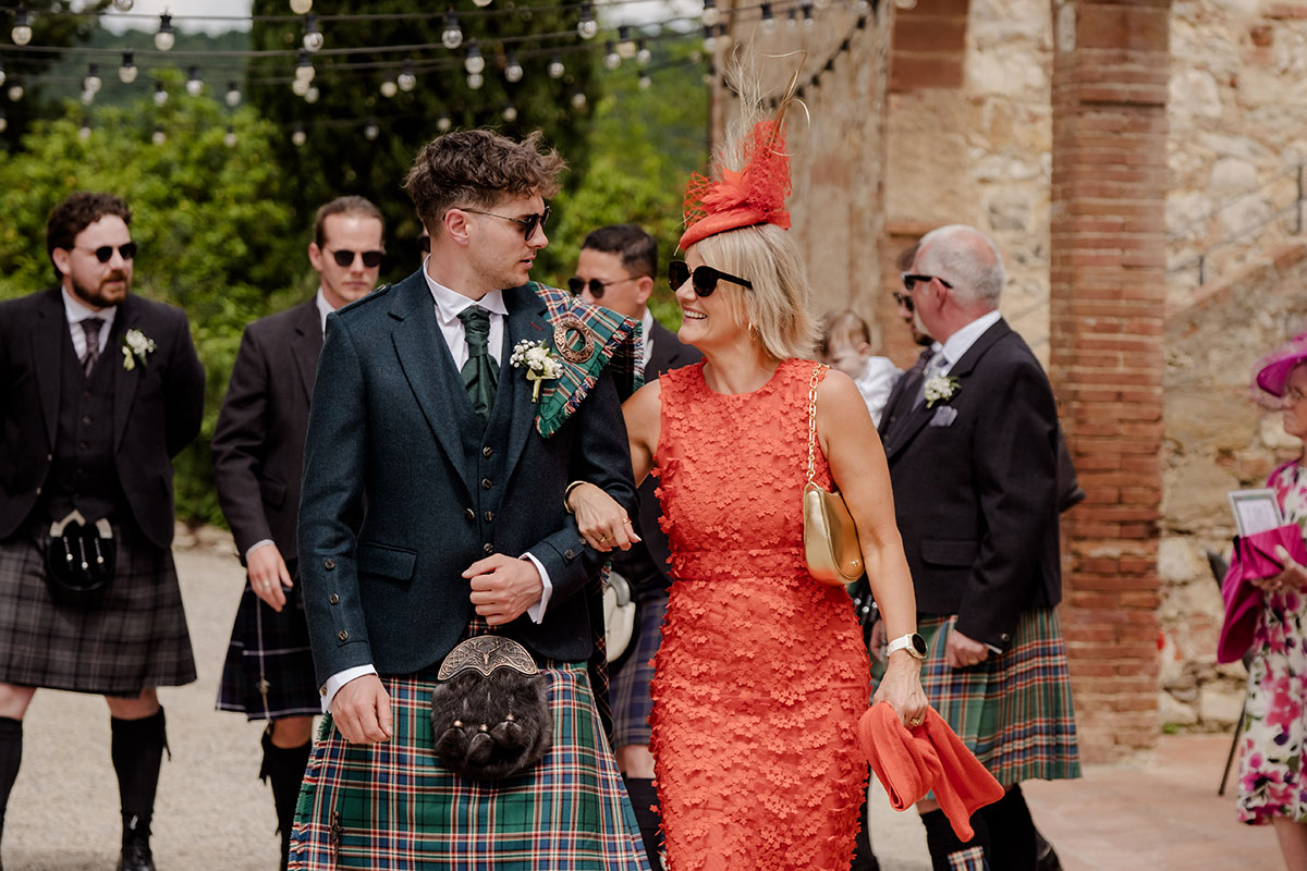 groom in blue, green and red tartan kilt outfit walks arm in arm with mum in orange midi dress and matching orange percher hat
