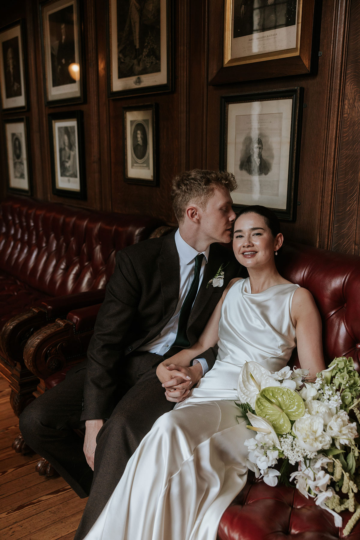 Groom kissing bride’s forehead as they sit on a red leather couch holding hands