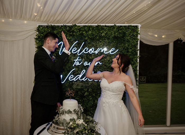 A bride and groom high five while standing behind their wedding cake and in front of a neon sign that says 'welcome to our wedding'