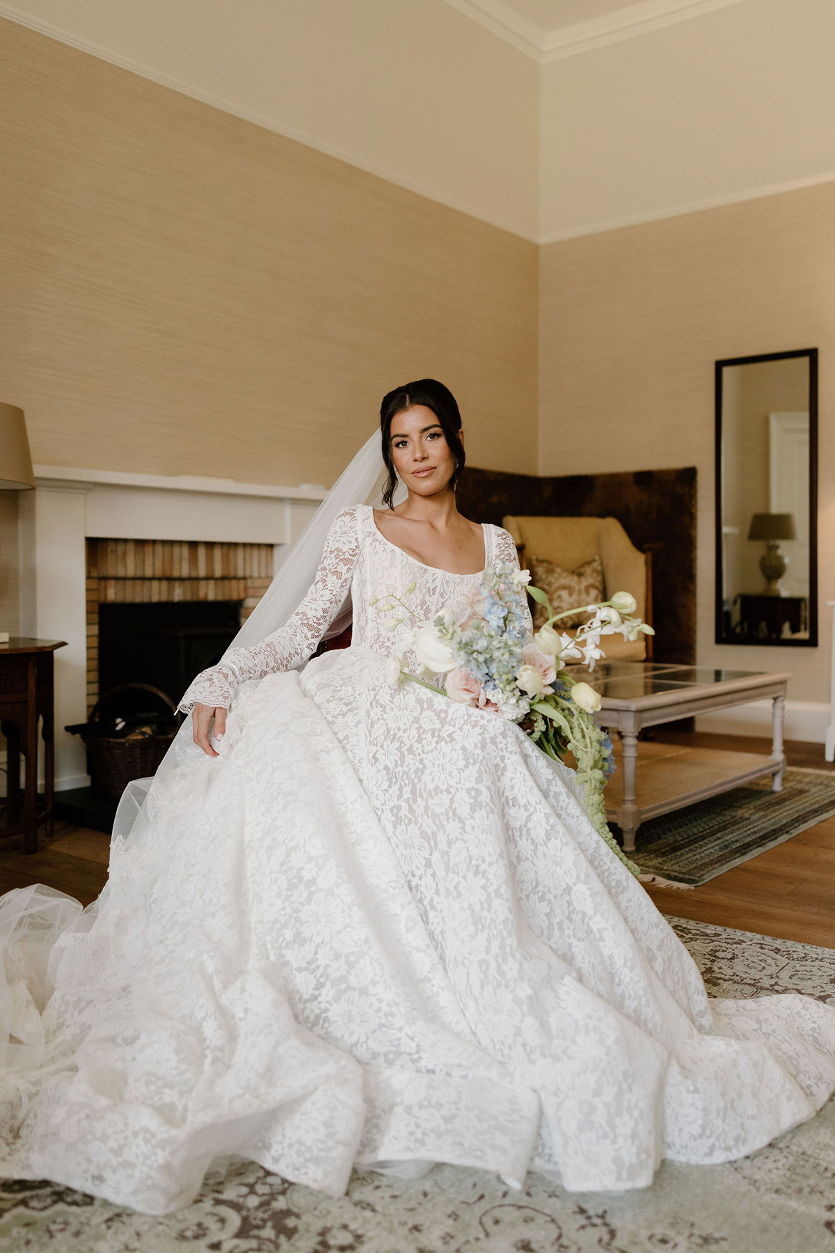 Bride in a lace wedding dress seated indoors holding a pastel bouquet