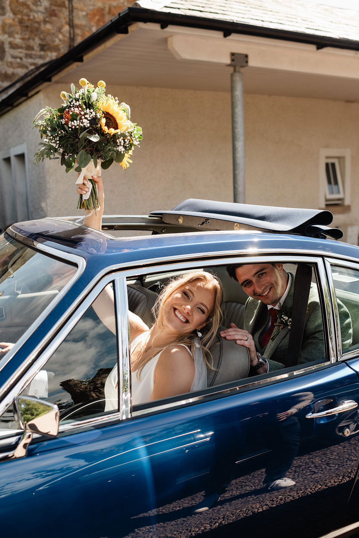 Newlyweds smiling from a classic blue wedding car as the bride holds a sunflower bouquet through the open sunroof.