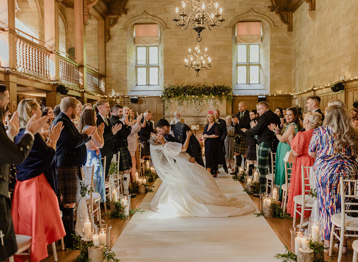 a groom and bride embrace and kiss on the white carpet aisle in the wood panelled ballroom at Achnagairn Castle as assembled wedding guests on either side cheer and clap