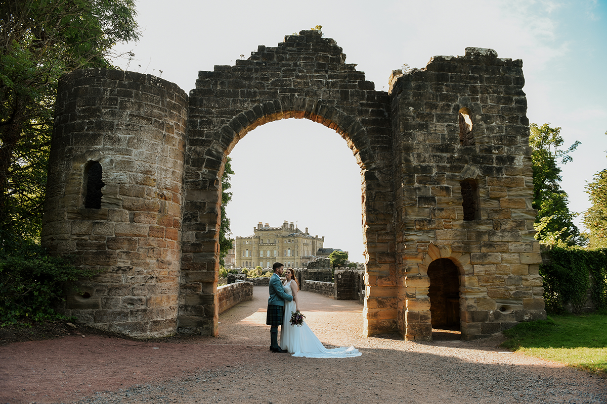 A couple stand beneath the stone archway at Culzean Castle, with the castle visible in the background.