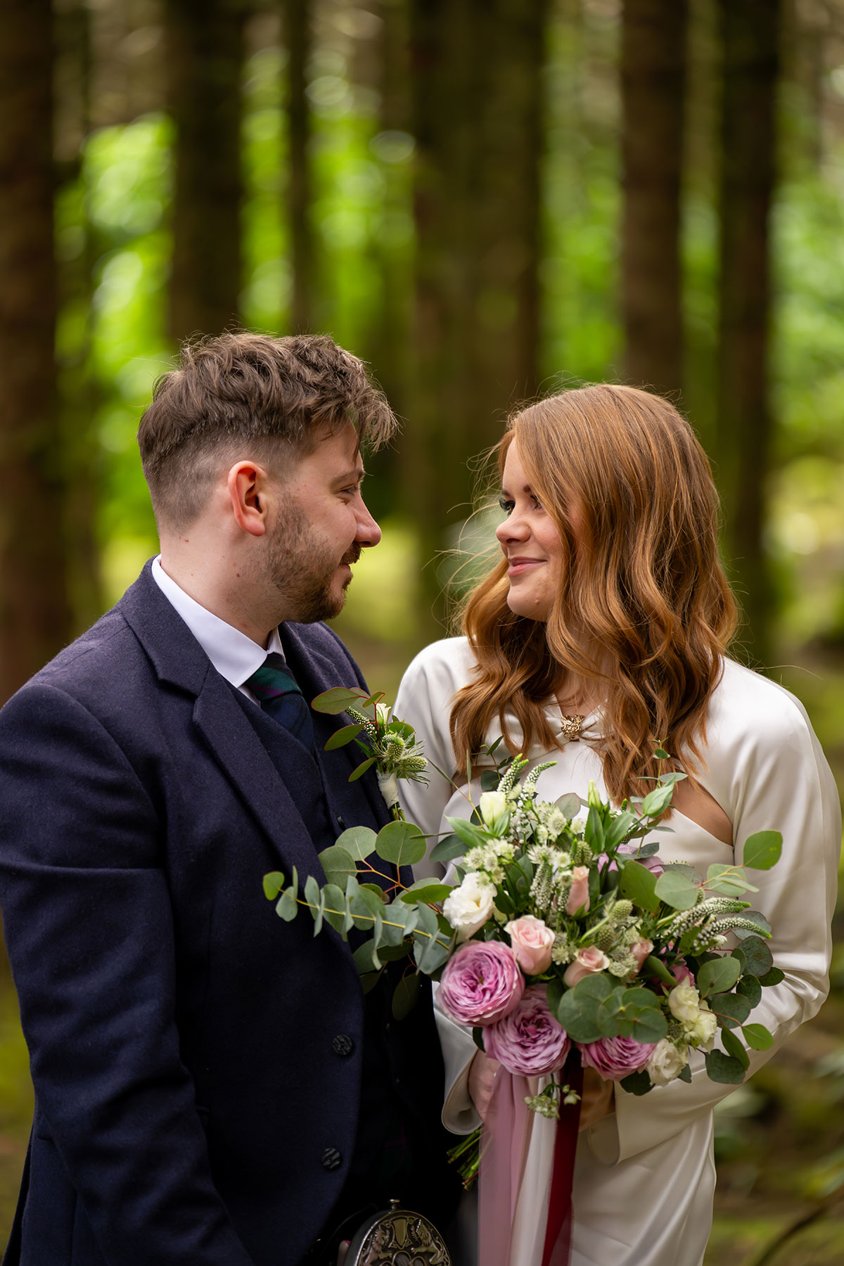 Bride and groom smiling at each other in a Scottish woodland after their Glencoe elopement, with the bride holding a bouquet of pink and white flowers