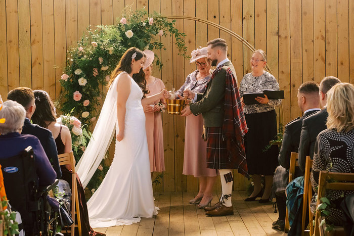 A bride and groom hold a gold quaich with their mothers during their wedding ceremony 
