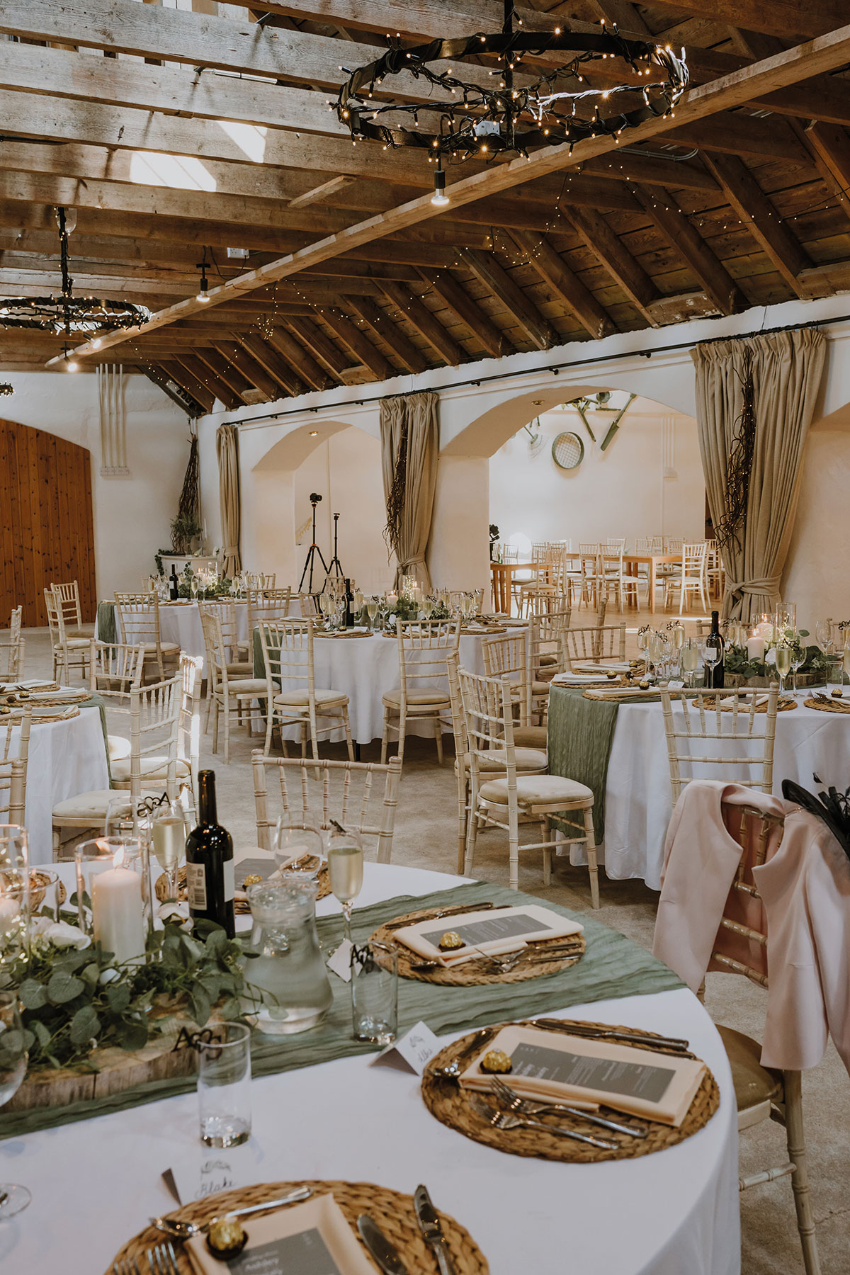 Wedding breakfast tables decorated with white linens, sage green runners, candles and greenery beneath exposed wooden beams and rustic chandeliers in Aswanley's dining room