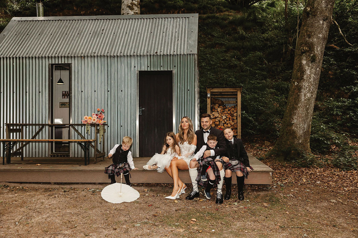 Bride and groom sitting with four children outside a small corrugated cabin in a woodland setting; the bride wears a short white dress and the groom and boys wear kilts