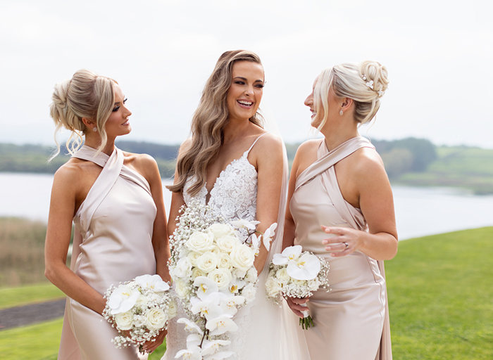 a bride laughing with two bridesmaids wearing oyster coloured cross-neck dresses. They are each carrying white orchid, white rose and gypsophila bouquets