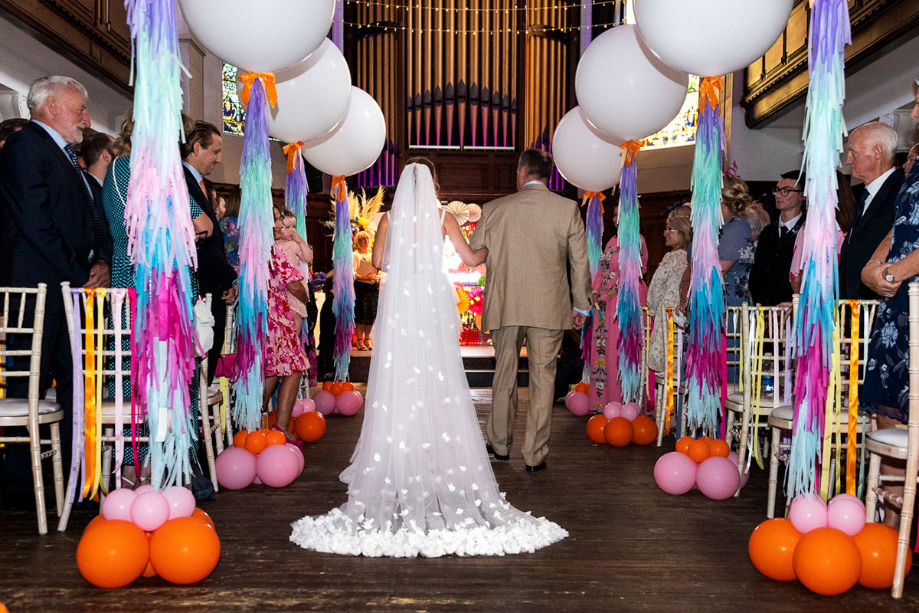Bride walks down aisle with her father. Aisle has been decorated with brightly coloured balloons
