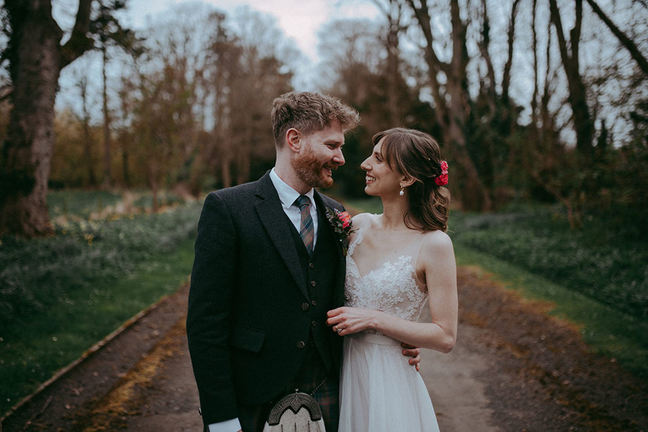 a bride and groom looking lovingly at one another. They are standing on a path in a garden setting with bare-branched trees behind them