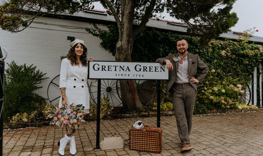 a bride and groom standing either side of a road sign that reads 'Gretna Green'. The bride is wearing a white blazer, a white beret and white boots and carrying a large pastel coloured bouquet. The groom is wearing a brown tweed suit with orange handkerchief tied around his neck