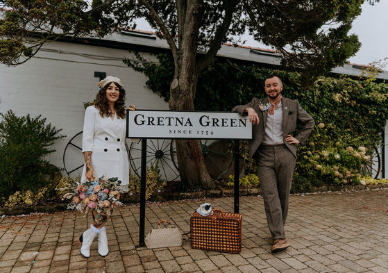 a bride and groom standing either side of a road sign that reads 'Gretna Green'. The bride is wearing a white blazer, a white beret and white boots and carrying a large pastel coloured bouquet. The groom is wearing a brown tweed suit with orange handkerchief tied around his neck