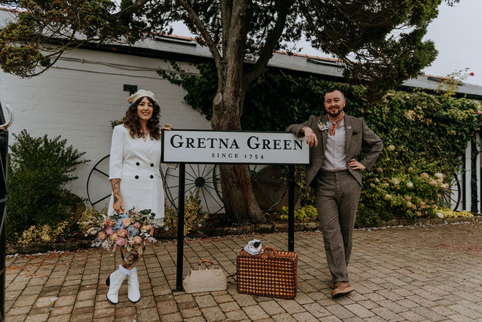 a bride and groom standing either side of a road sign that reads 'Gretna Green'. The bride is wearing a white blazer, a white beret and white boots and carrying a large pastel coloured bouquet. The groom is wearing a brown tweed suit with orange handkerchief tied around his neck