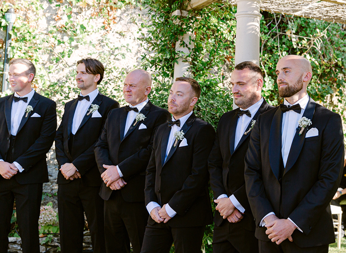 a line up of men standing in a garden wearing smart black tuxedo suits with bow ties 