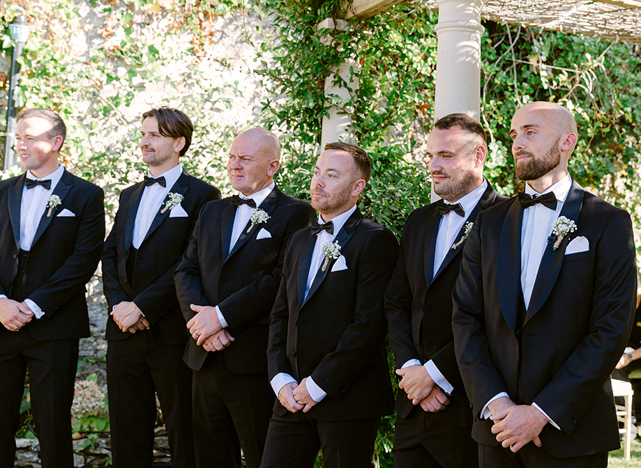 a line up of men standing in a garden wearing smart black tuxedo suits with bow ties