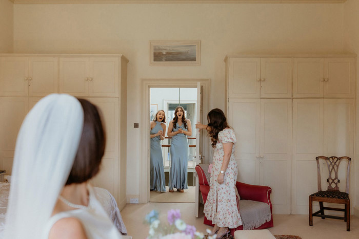 two bridesmaids wearing blue Six Stories bridesmaid dresses looking through an open door towards a bride in a room with ivory cupboards