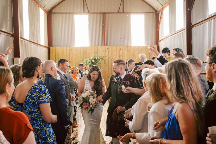 A bride and groom leaving their ceremony in a barn as the guests throw confetti over them
