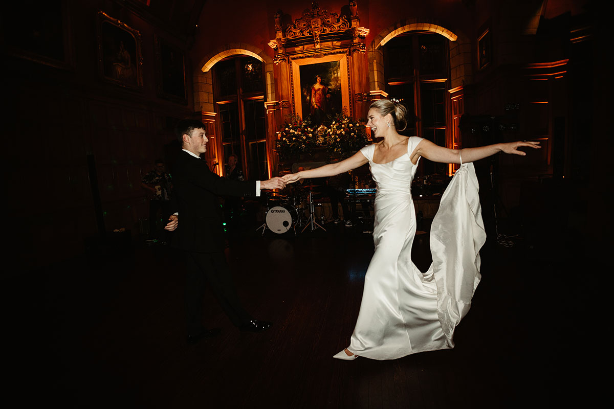 Bride and groom dancing in the Minstrels’ Hall, holding hands under warm amber lighting.