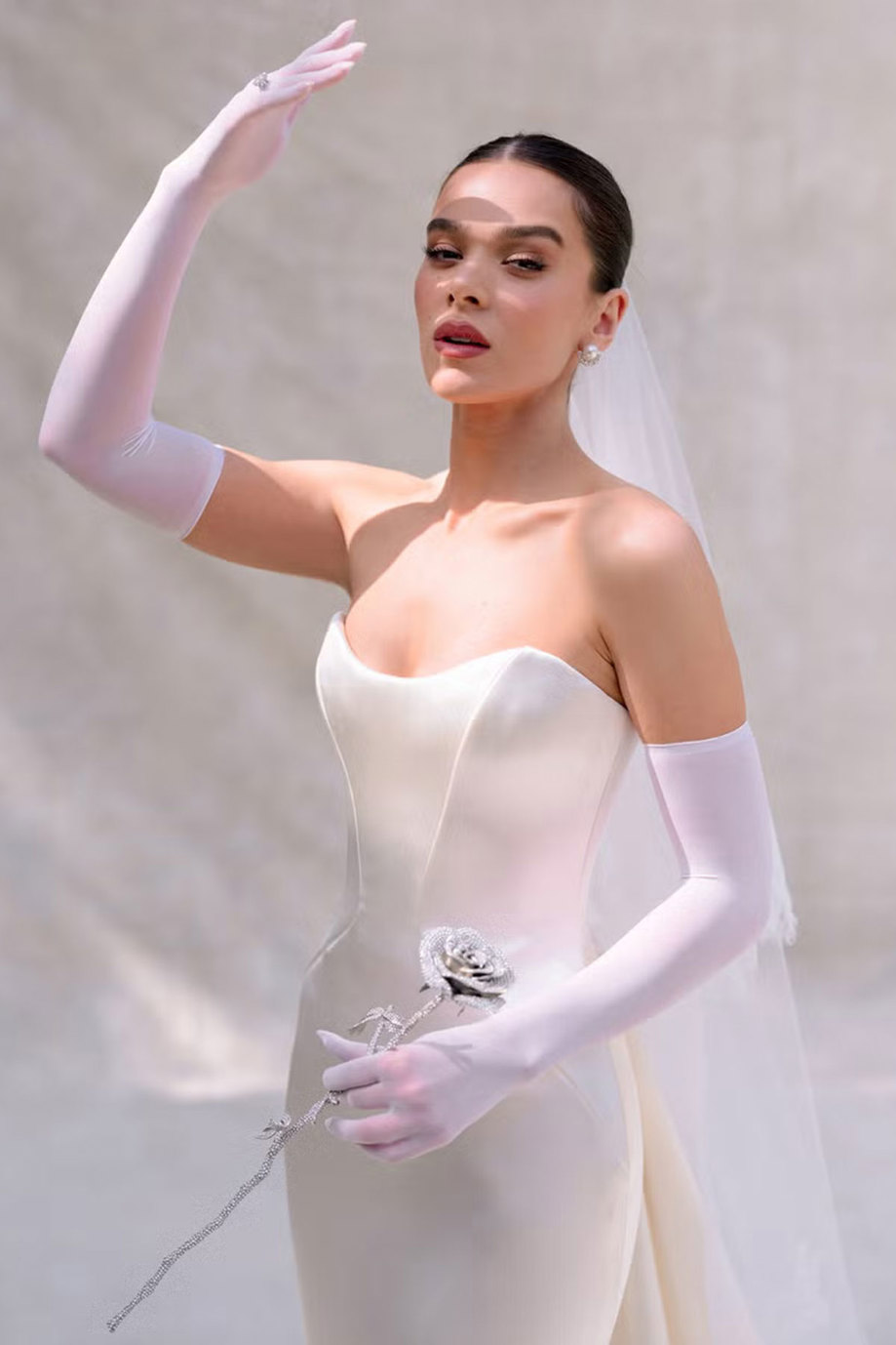 brunette bride in a silky smooth strapless wedding dress looks up at the sun, holding her sheer elbow-length gloved hand up in front of her face while the other holds a single glittery fake rose