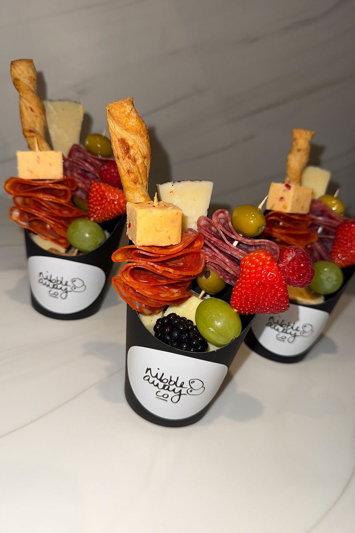 Three black branded graze cups filled with cheese cubes, cured meats, olives, grapes, berries and breadsticks, arranged upright on a marble surface