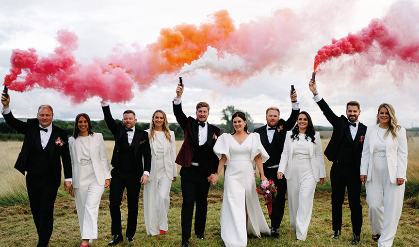 a group of people wearing wedding attire holding red, pink and orange smoke bombs walking on grass