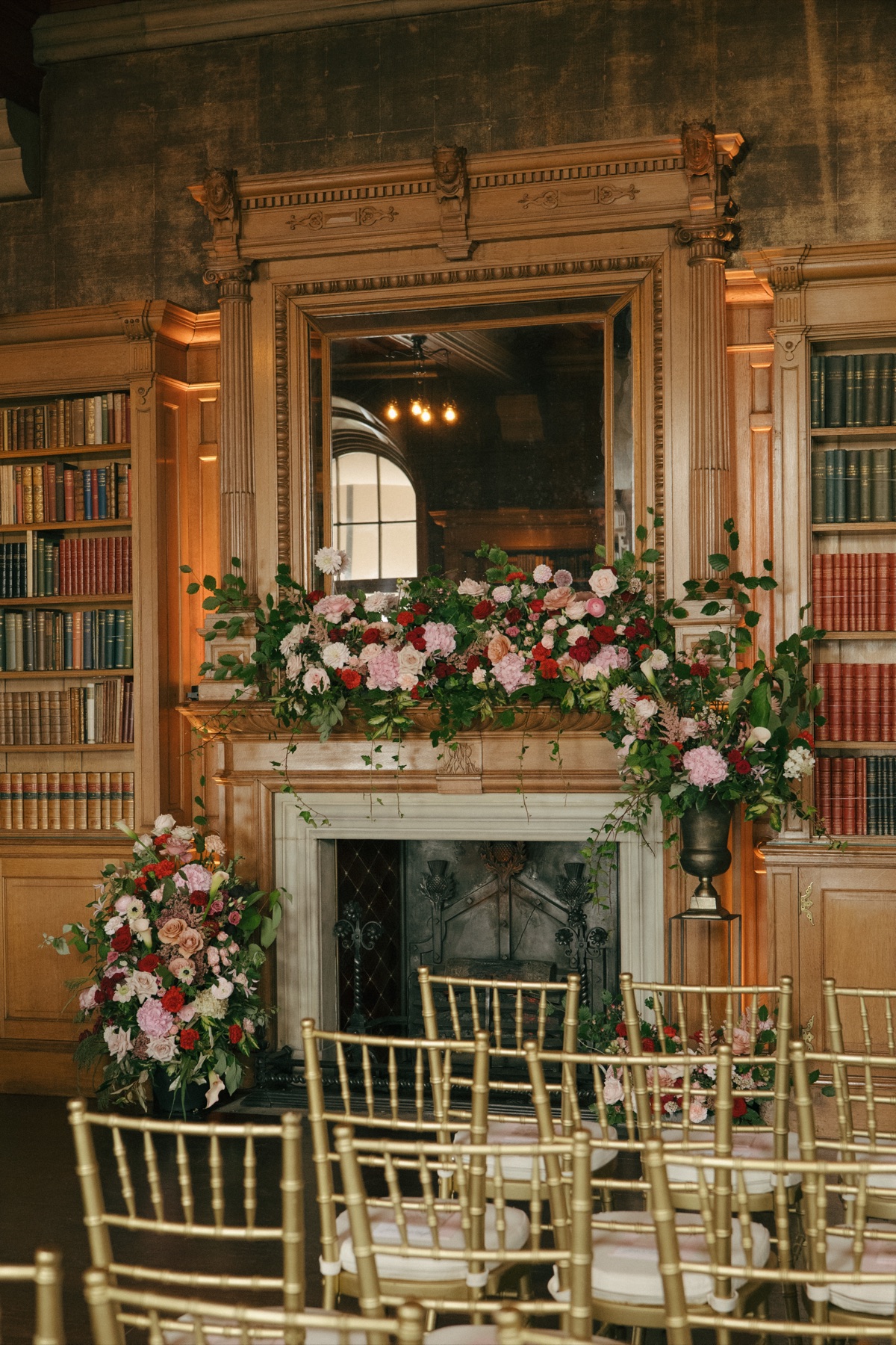Pink, red and cream wedding flowers decorate the fireplace in Barnbougle Castle's library, with gold chairs arranged for the ceremony.