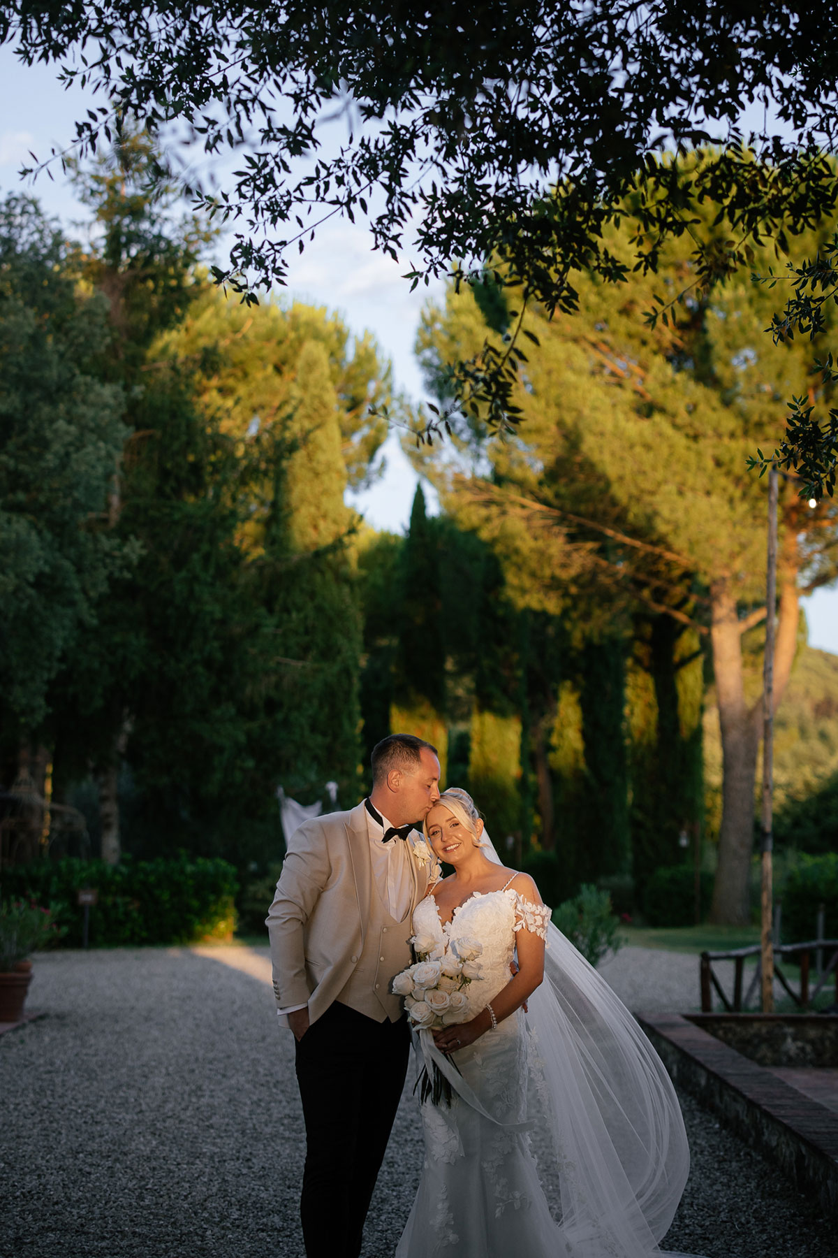Bride and groom standing outdoors at sunset, with the groom kissing the bride’s head as she smiles and holds a bouquet of white roses, surrounded by tall trees