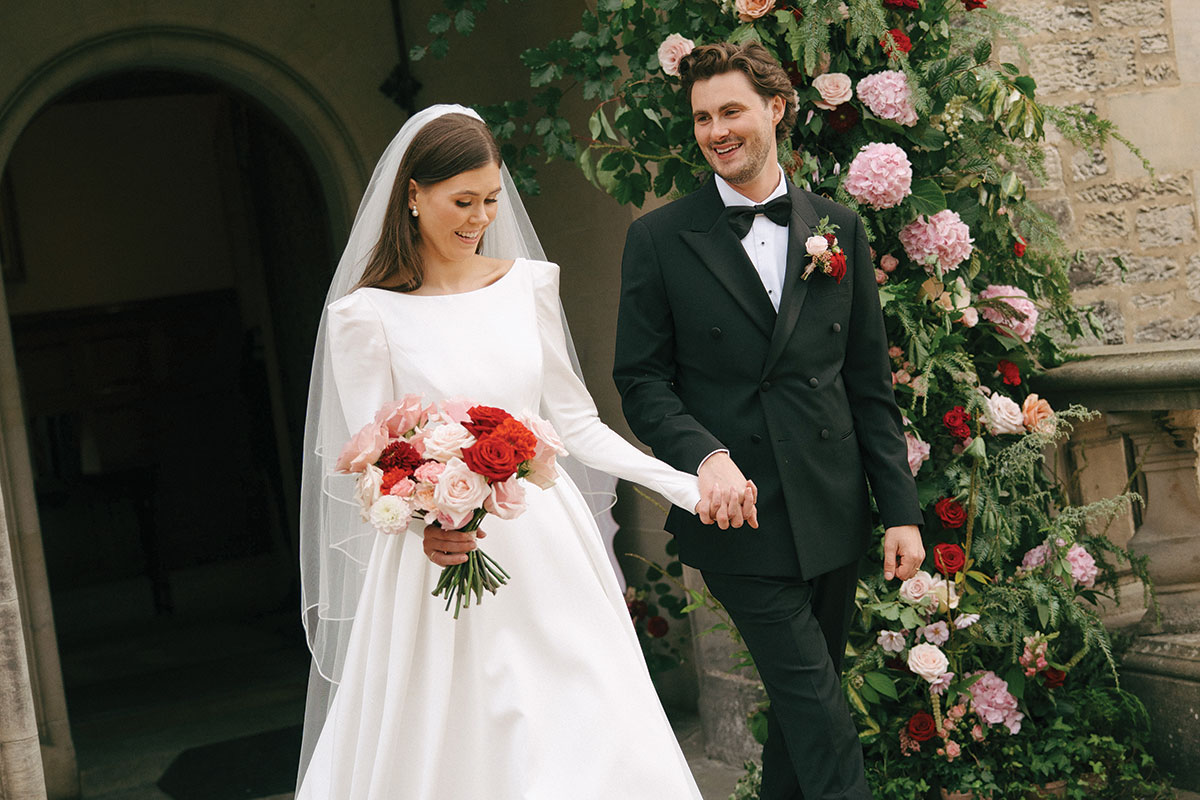 Bride Nicole and groom Ryan walk hand in hand outside Barnbougle Castle in Edinburgh, beside a floral display of pink and red roses.