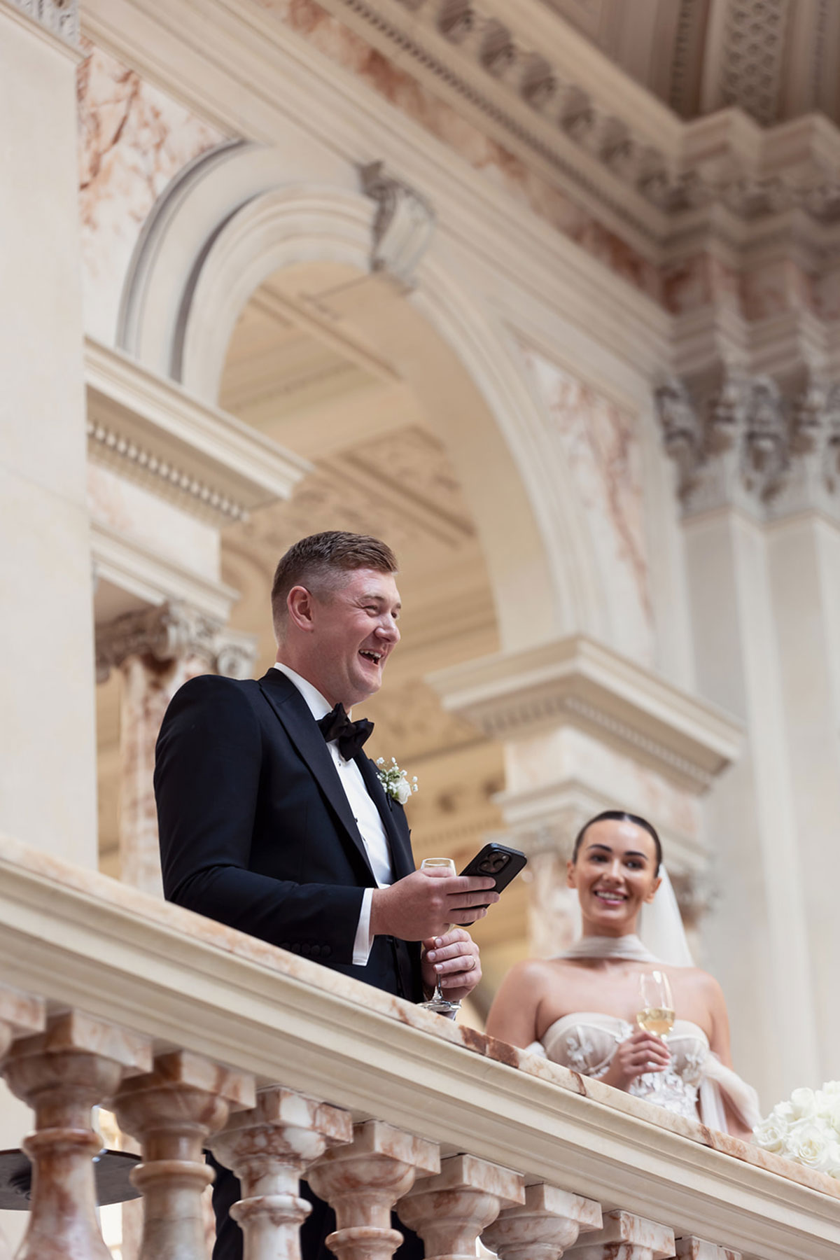 A bride holding a glass of champagne and smiling at a person in a tuxedo.