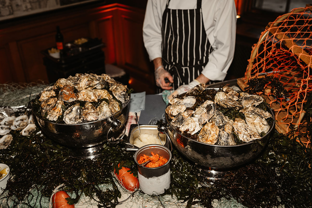 Large silver bowls filled with oysters on ice and seaweed, with a chef preparing them behind the display.