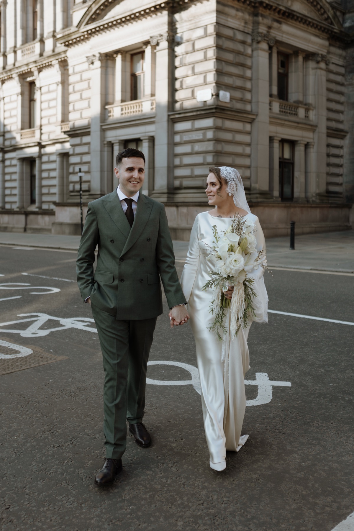 Newlyweds walking hand in hand through Glasgow city centre, with the bride carrying a cascading white bouquet.