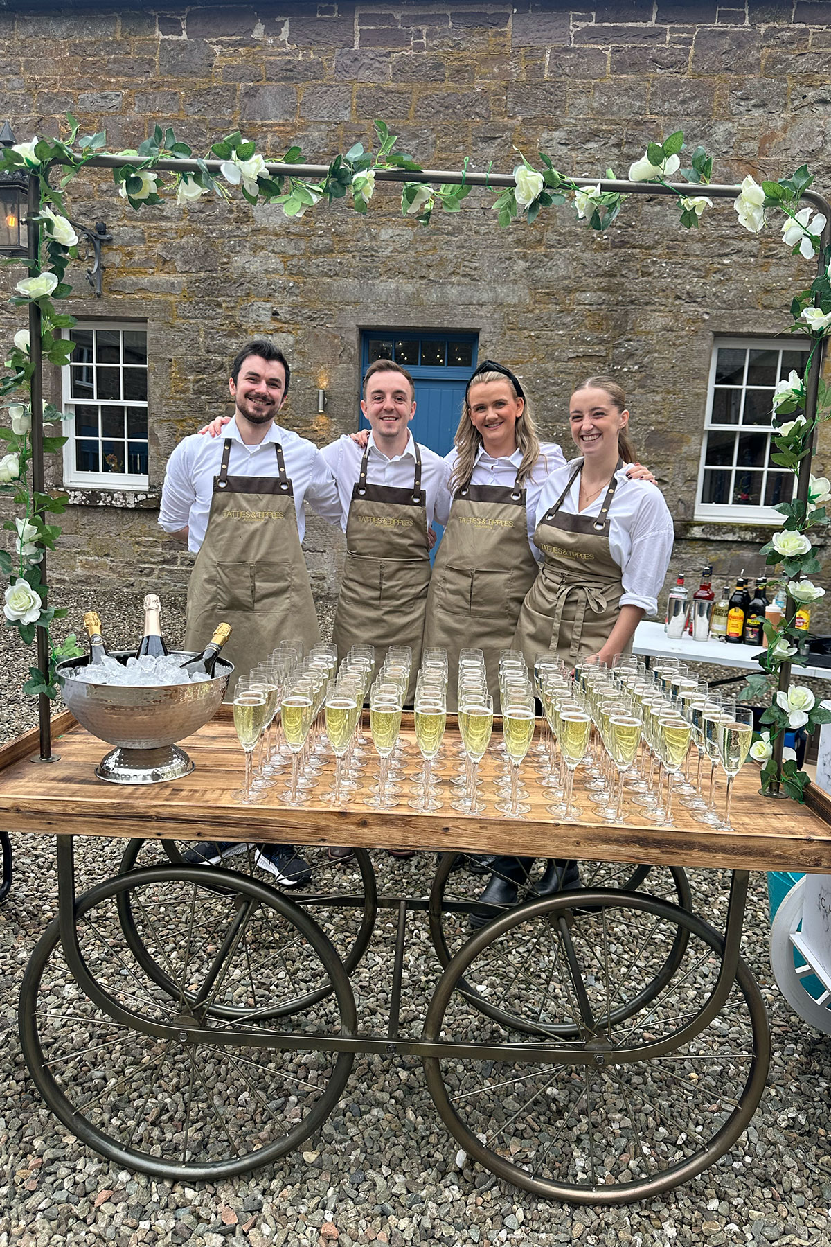 The Tatties and Tipples bar team standing behind a rustic drinks cart filled with chilled champagne glasses at a wedding venue