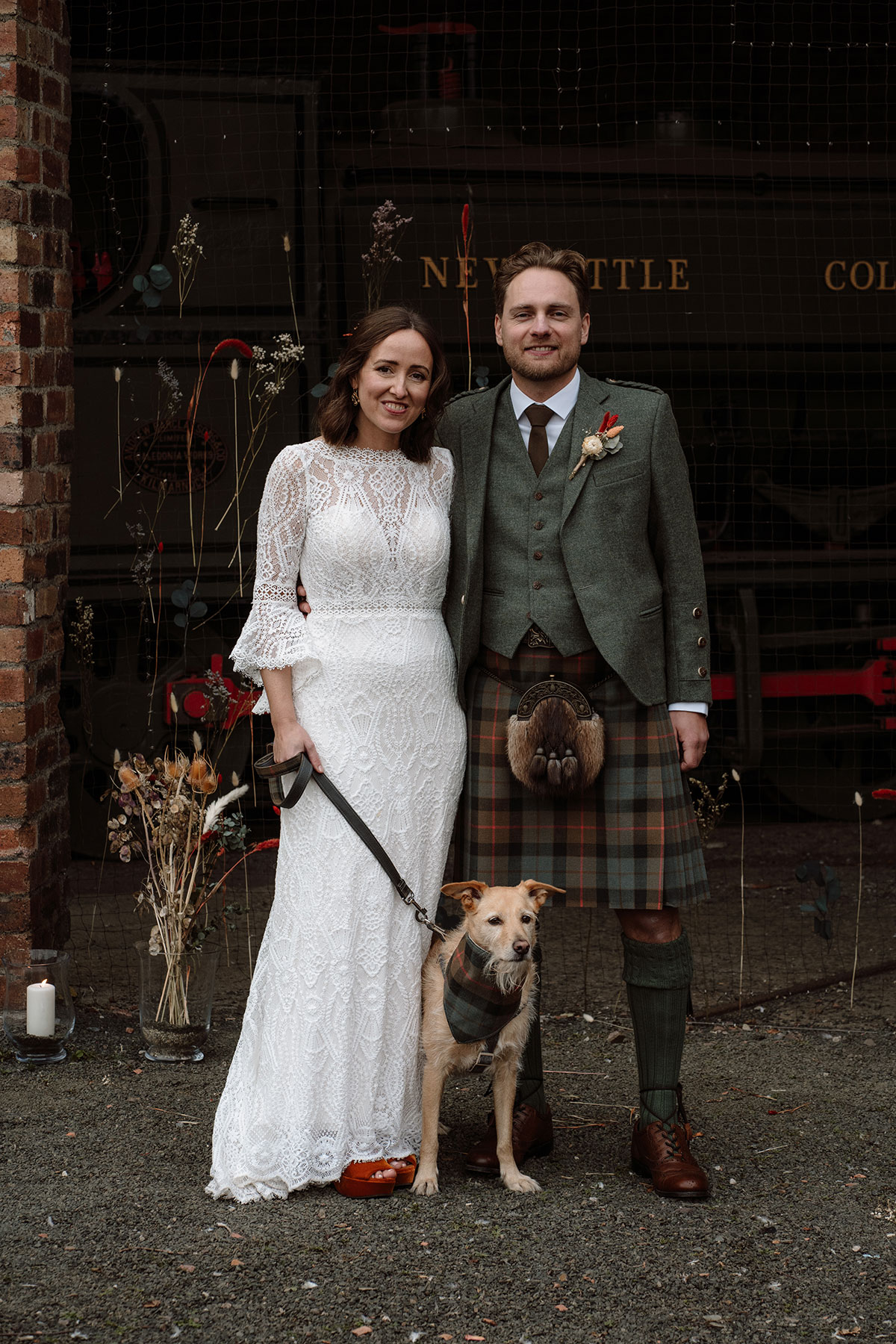 Bride and groom pose with their dog outside the venue, with dried floral arrangements and industrial heritage features in the background