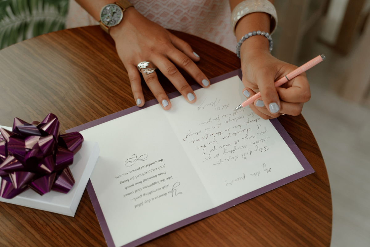 Person writing a handwritten message inside a wedding card beside a wrapped gift.