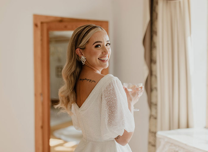a smiling bride with tattoo on her back holding a champagne coupe looks over her shoulder. She wears an Eva Lendel Rosie wedding dress and pearl earrings from Untamed Petals.