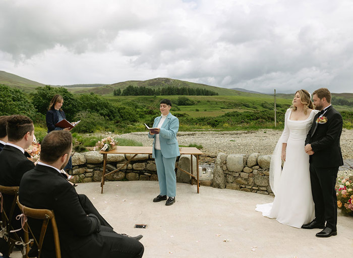 a person wearing a blue suit reading from a book during a wedding ceremony outdoors. The bride and groom stand to one side watching on and an officiant stands on the other. There is a wall, stony ground and green fields in the background