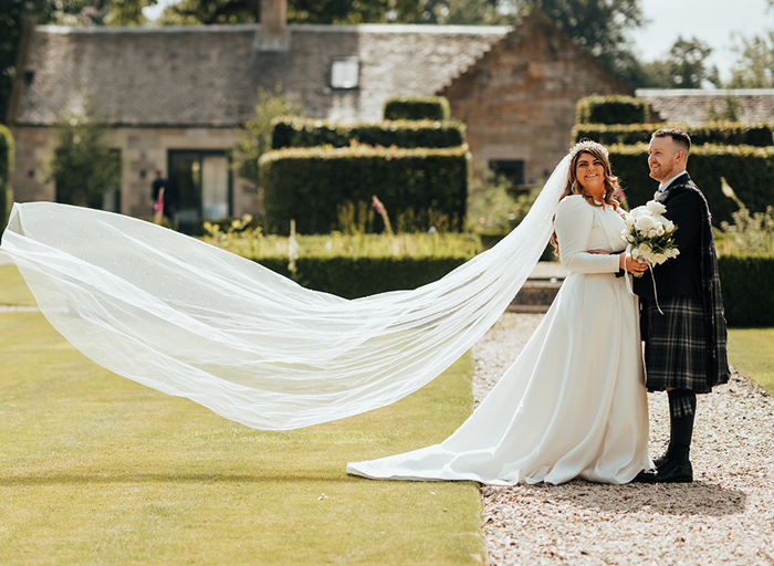 A man in a dark kilt holding hands with a bride in a long sleeved wedding dress as her veil floats behind her