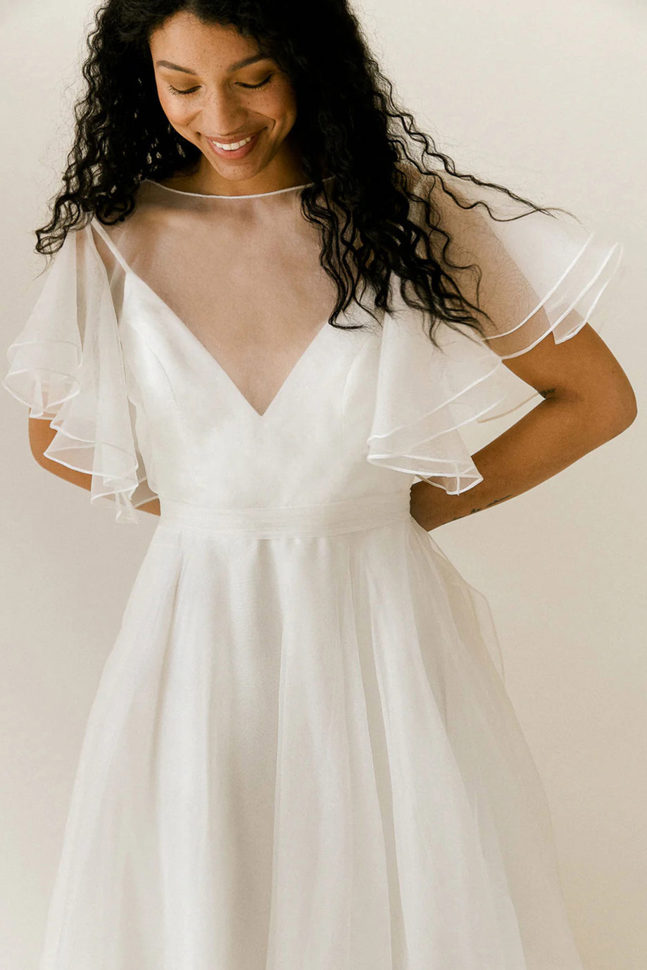 curly dark-haired model wears v-neck white wedding dress with sheer layer over bodice providing fluttery sleeves