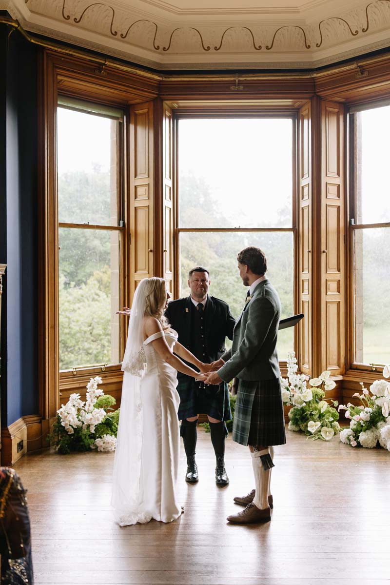 Lia and Louis hold hands during their wedding ceremony in the flower-filled library at Cambo House in Fife.