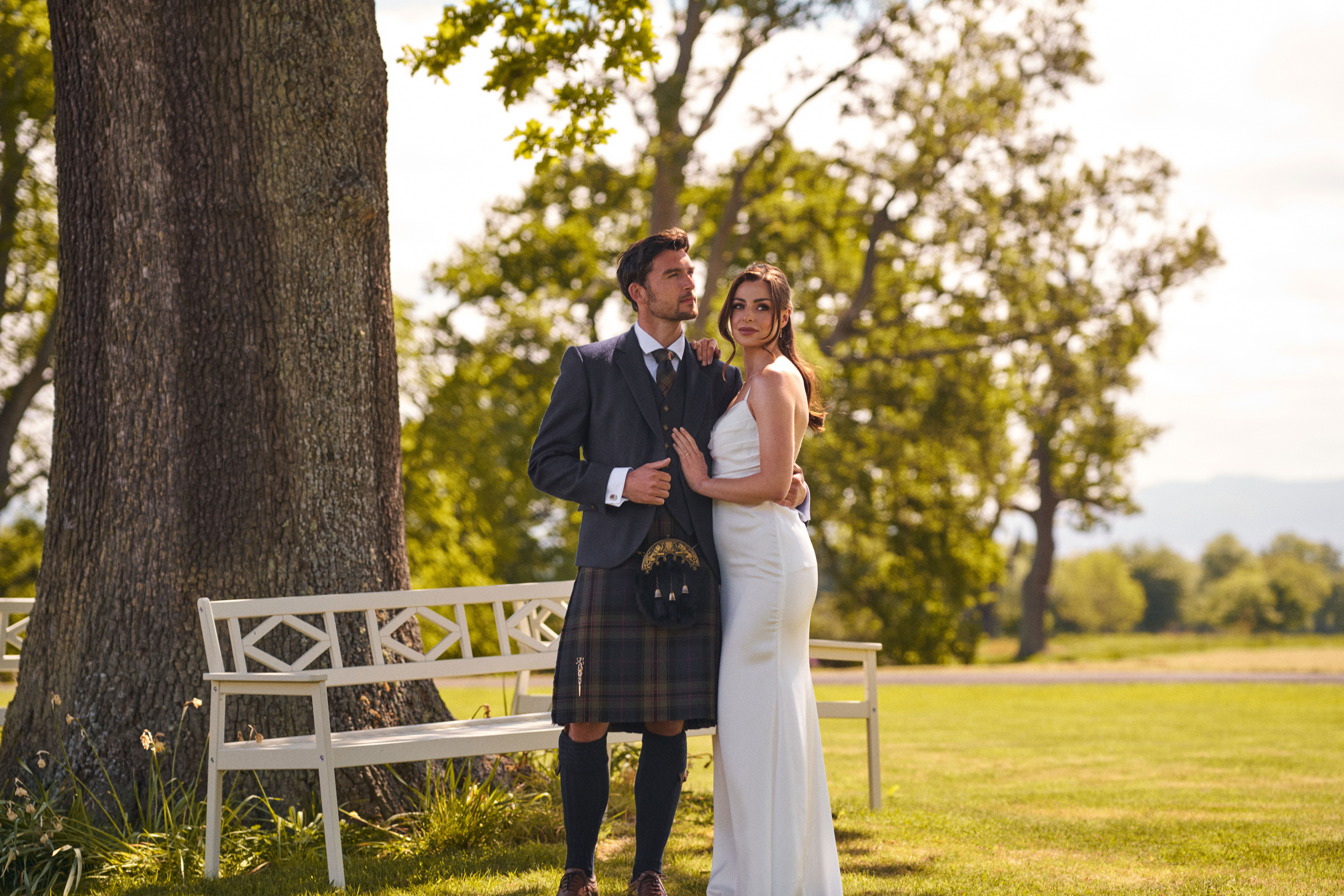bride and groom stand in front of white bench with green grass in the background at dundas castle while wearing mccalls highlandwear's new noble print tartan kilt