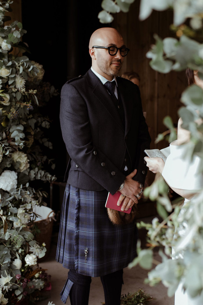 groom in blue, grey and white tartan kilt outfit stands listening as bride reads from little red booklet of vows at an altar surrounded by white and green florals